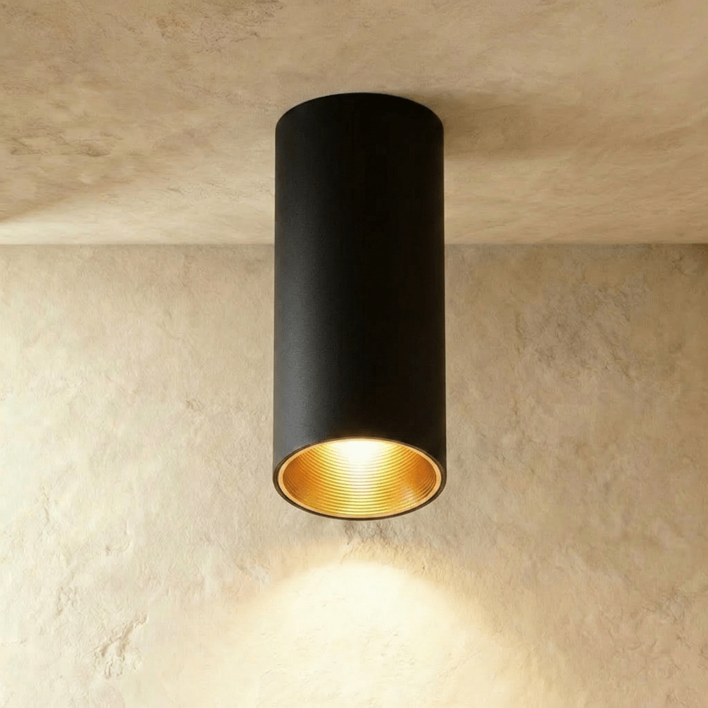 Clérachic surface downlight in black aluminium with a gold-tone baffle, casting warm light onto a plaster ceiling corner.