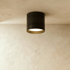 Clérachic larger black surface-mounted downlight with two fine grooves and a gold-toned inner rim, lit on a plaster ceiling.