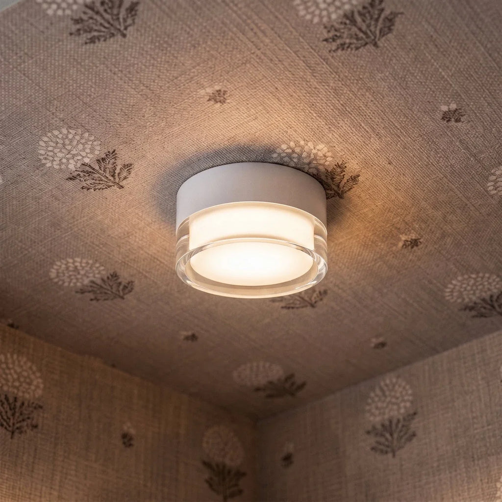 Clérachic white round flush ceiling light with a clear acrylic rim, lit warmly against a patterned fabric-covered ceiling.