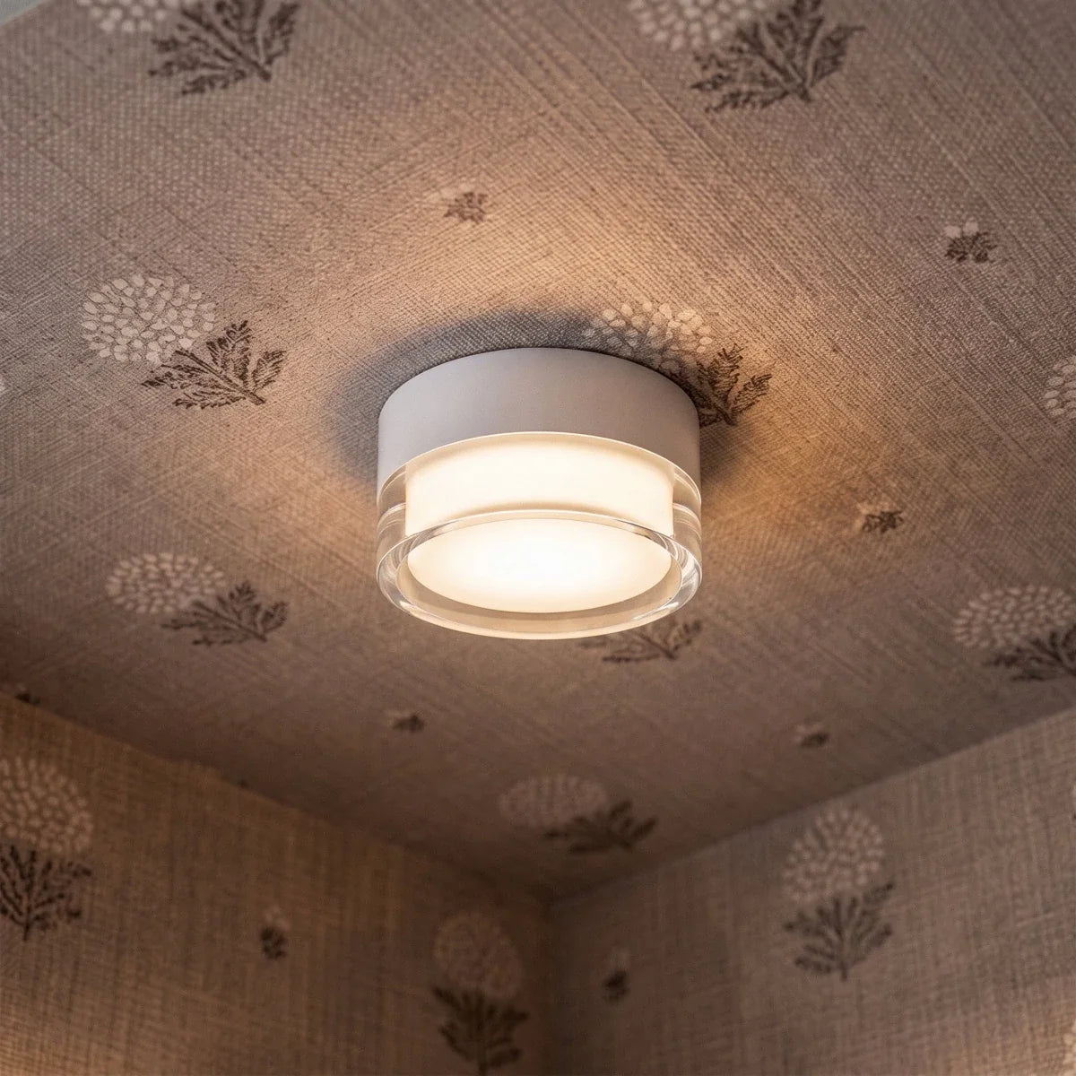 Clérachic white round flush ceiling light with a clear acrylic rim, lit warmly against a patterned fabric-covered ceiling.