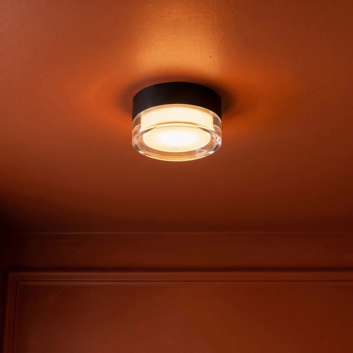 Clérachic black round ceiling light with a clear acrylic edge casting a warm amber glow across a terracotta-painted ceiling.