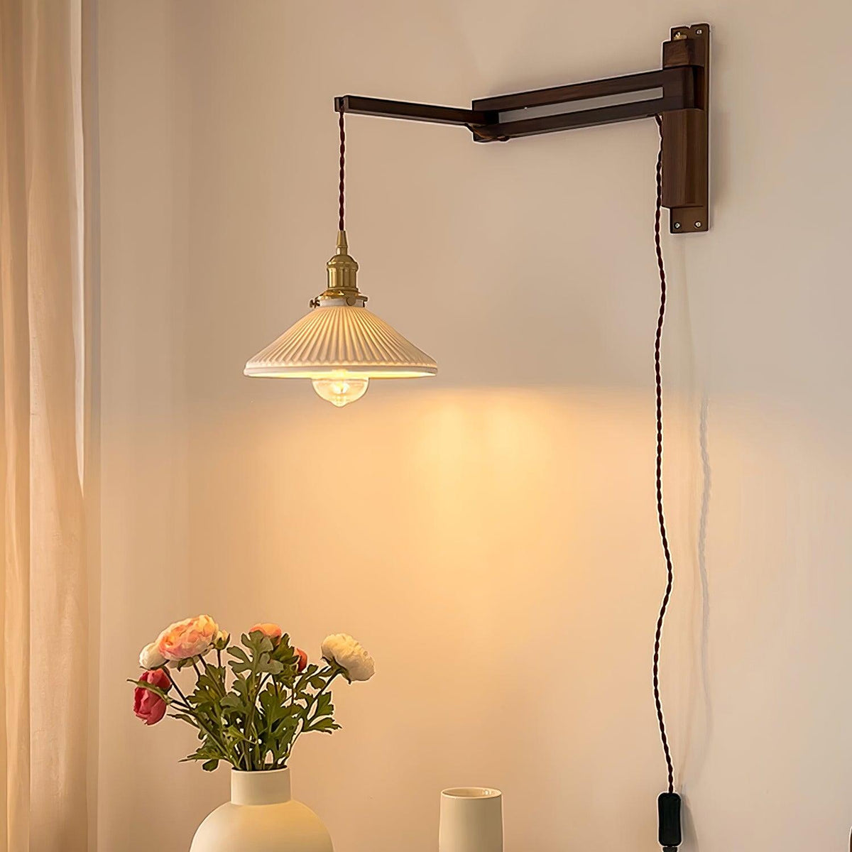Pleated white shade glowing above a vase of flowers, on a Clérachic plug-in wall light with a long dark wood swing arm and a twisted flex.