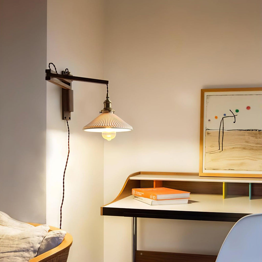 A Clérachic swing-arm wall light lit beside a wooden desk and framed print, its pleated shade hanging from a slim dark wood arm.