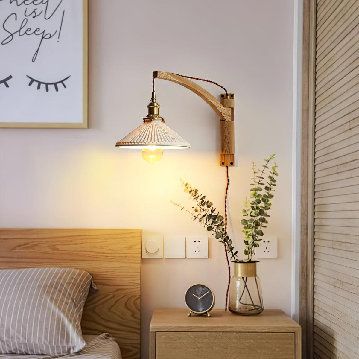 Above a bedside table and wooden headboard, a Clérachic wall light with a curved light-wood arm and a pleated ceramic shade lit warm.