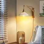 Warm light from a Clérachic wall light with a curved wood arm, beside a rattan lantern and a window blind in a bedroom corner.