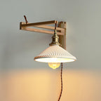 Close view of the pleated white ceramic shade and brass fitting on this Clérachic wall light, lit warm against a pale wall.