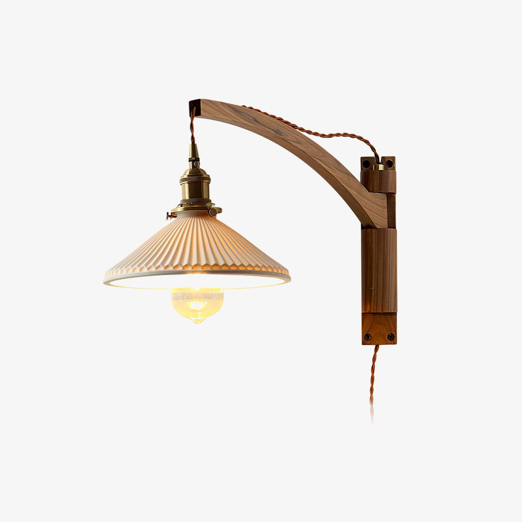 Plain backdrop view of this Clérachic wall light with a curved dark wood arm, brass socket, pleated white shade and twisted brown flex.
