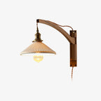 Plain backdrop view of this Clérachic wall light with a curved dark wood arm, brass socket, pleated white shade and twisted brown flex.