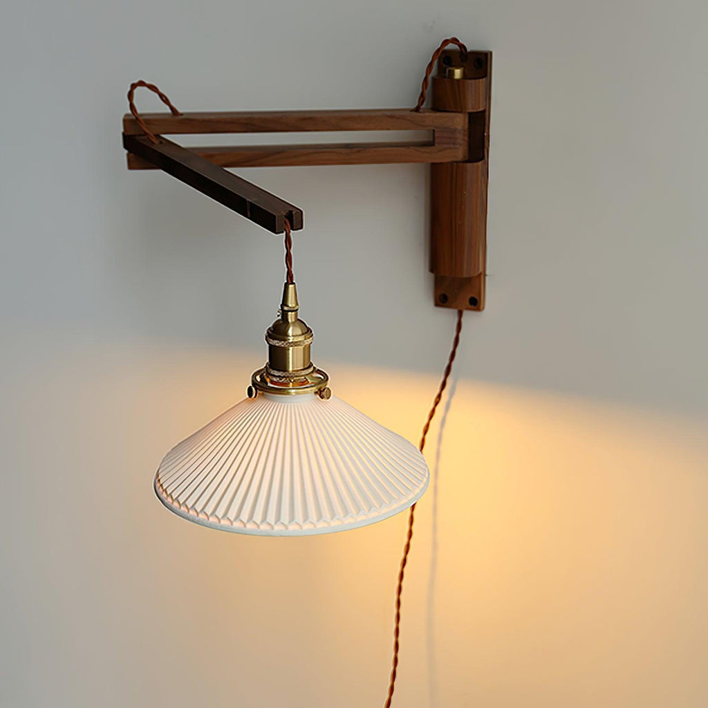 The folding wood arm and pleated ceramic shade of this Clérachic wall light, lit warm on a pale grey wall with the flex trailing down.