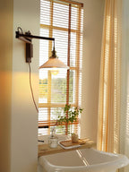 A Clérachic wall light beside a tall window with slatted blinds, the pleated shade on its swing arm above a bathtub and a small plant.