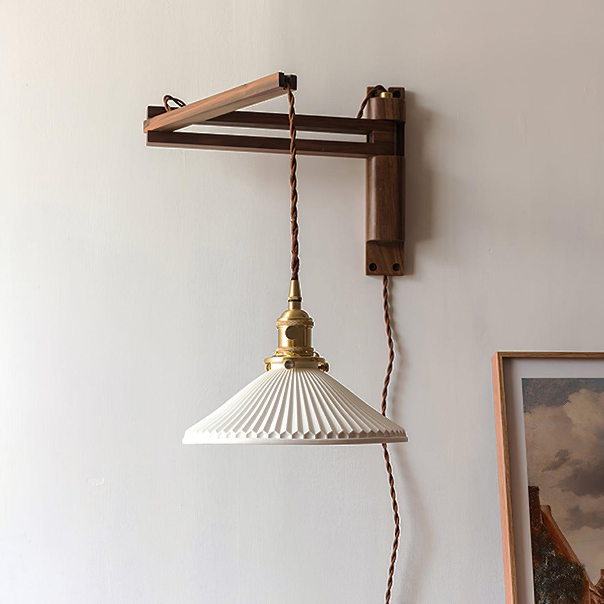 Pleated white shade hanging low from the walnut arm of this Clérachic wall light, with a framed landscape print at the edge of the shot.