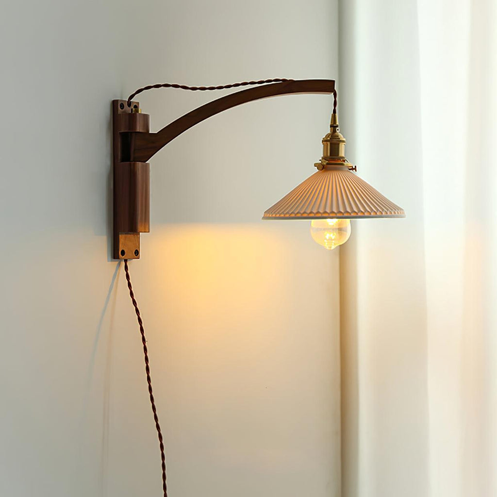 Curved walnut arm and pleated shade on this Clérachic wall light, with a thin brown flex and a white curtain behind.