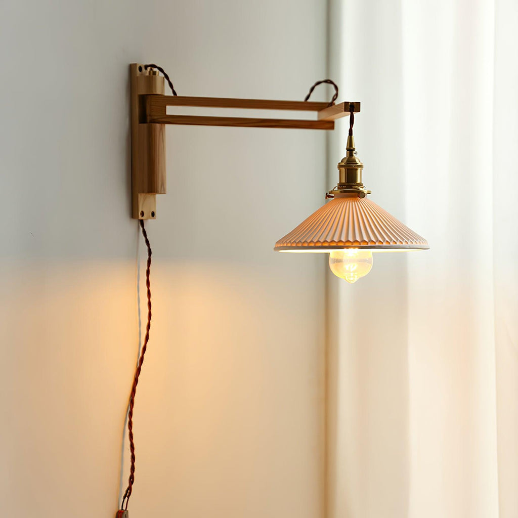 Square wood arm and brass socket of this Clérachic wall light, the pleated shade glowing against a grey wall beside a white curtain.