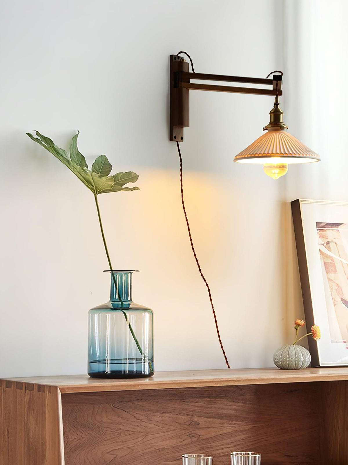 A Clérachic wall light in a bright room, a tall plant in a blue glass vase on a walnut sideboard below its pleated shade.