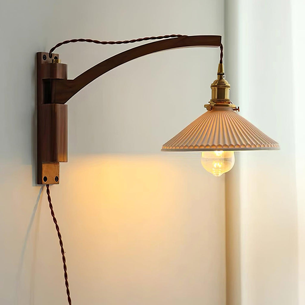 Dark walnut curved arm and back plate on this Clérachic wall light, its pleated shade lit warm and the flex hanging free.