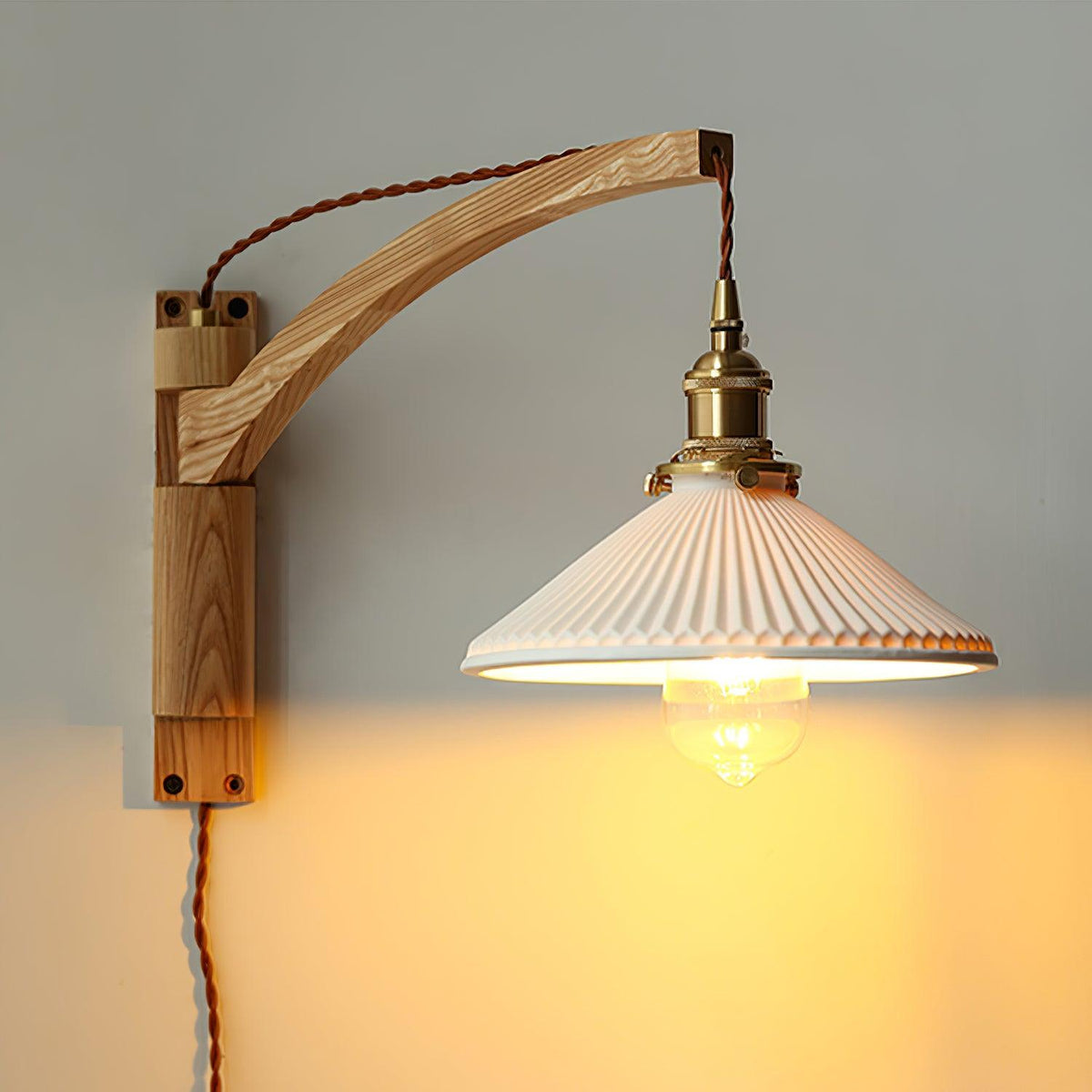 A Clérachic wall light with a light curved wood arm, the pleated white shade lit warm over a grey wall with screw heads on the back plate.
