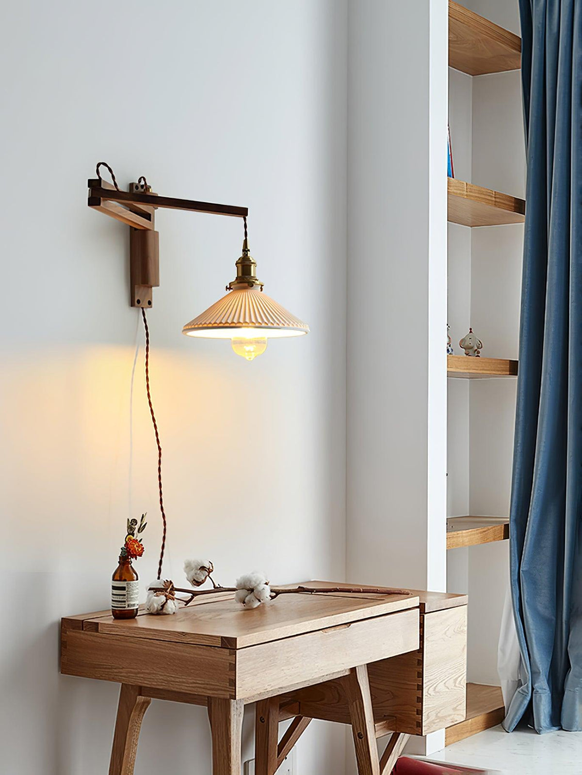 Seen from across a small wooden desk, this Clérachic wall light hangs its pleated shade from a dark wood arm beside a blue curtain.