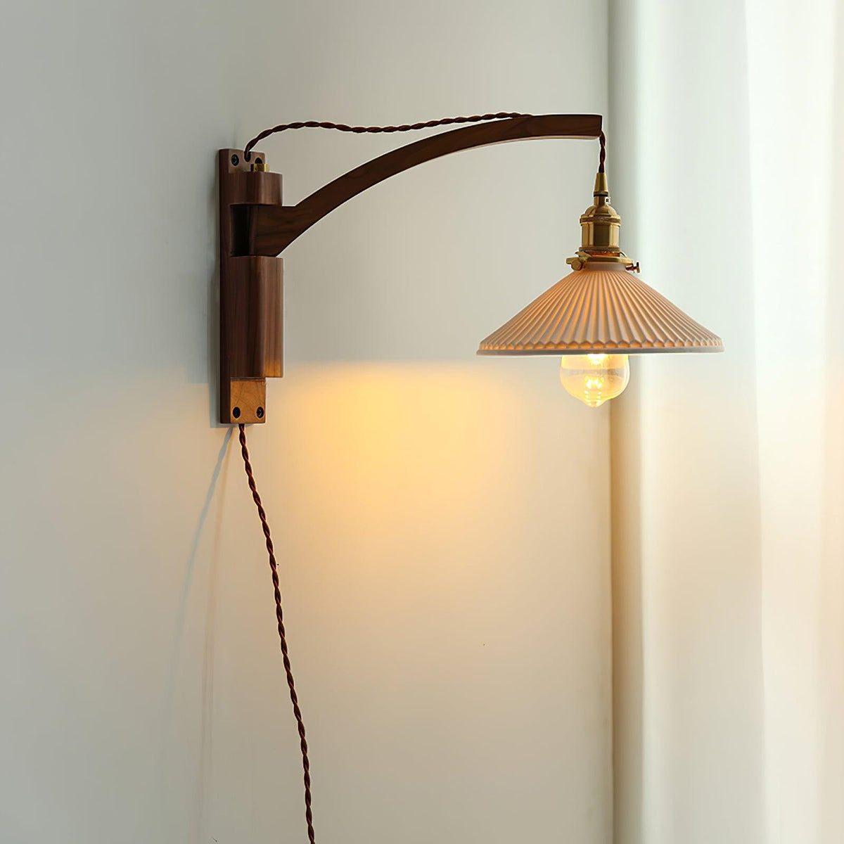 A Clérachic wall light with a curved dark wood arm in a pale corner, the pleated shade lit warm with a white curtain beside it.
