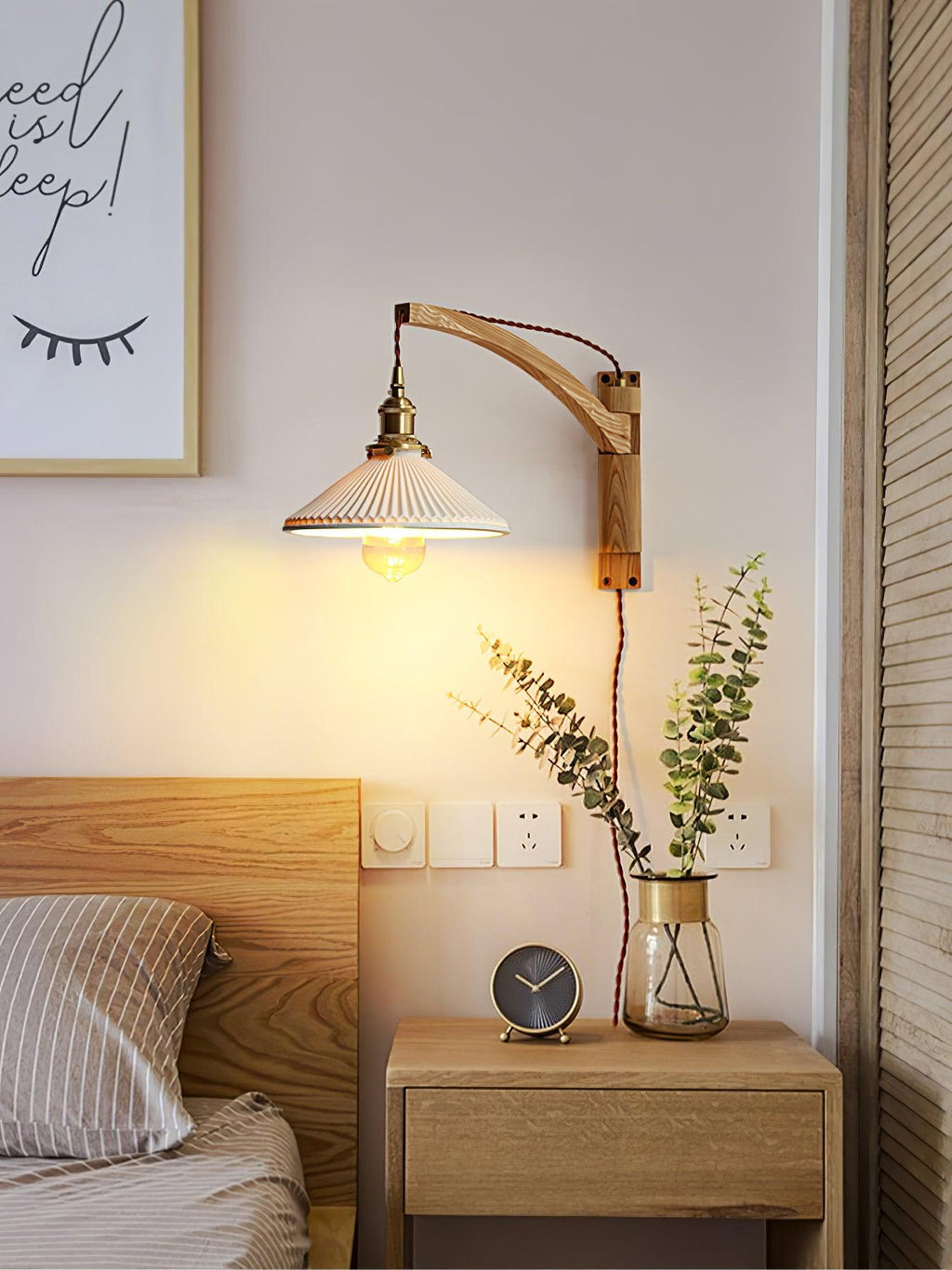 In a bedroom, a Clérachic wall light with a curved light-wood arm hangs above a nightstand with a clock and a glass vase of eucalyptus.