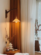 By a tall window with an amber curtain, a Clérachic wall light shows its pleated shade lit softly above a bedside table.