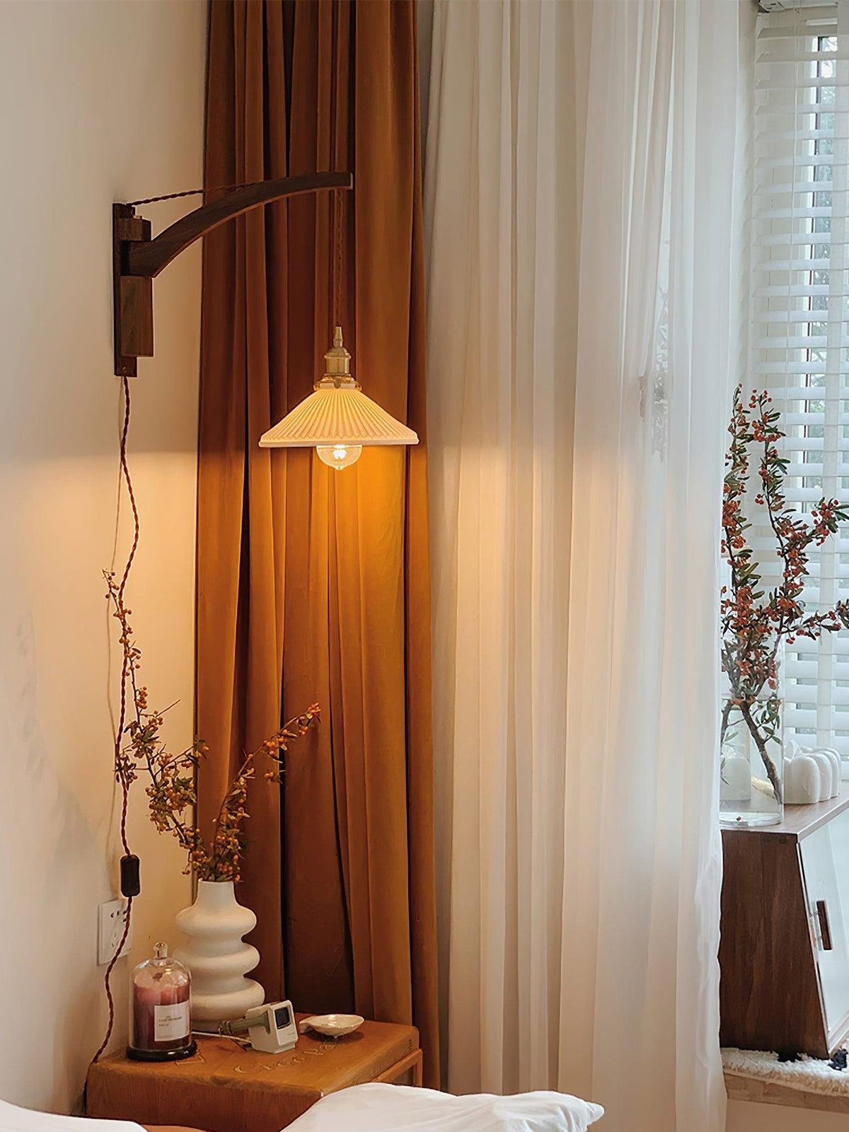 By a tall window with an amber curtain, a Clérachic wall light shows its pleated shade lit softly above a bedside table.