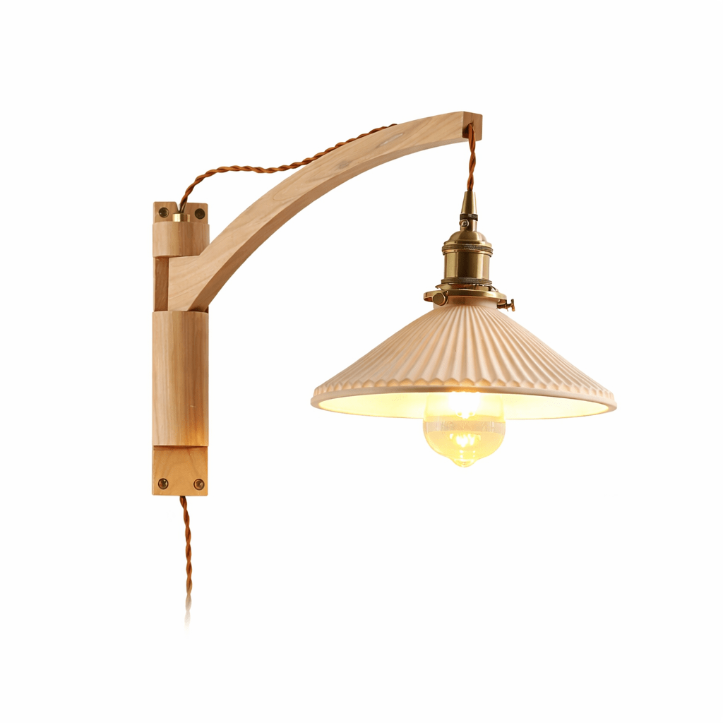 On white, this Clérachic wall light has a gently curved light-wood arm and a pleated shade with a warm bulb glowing under it.