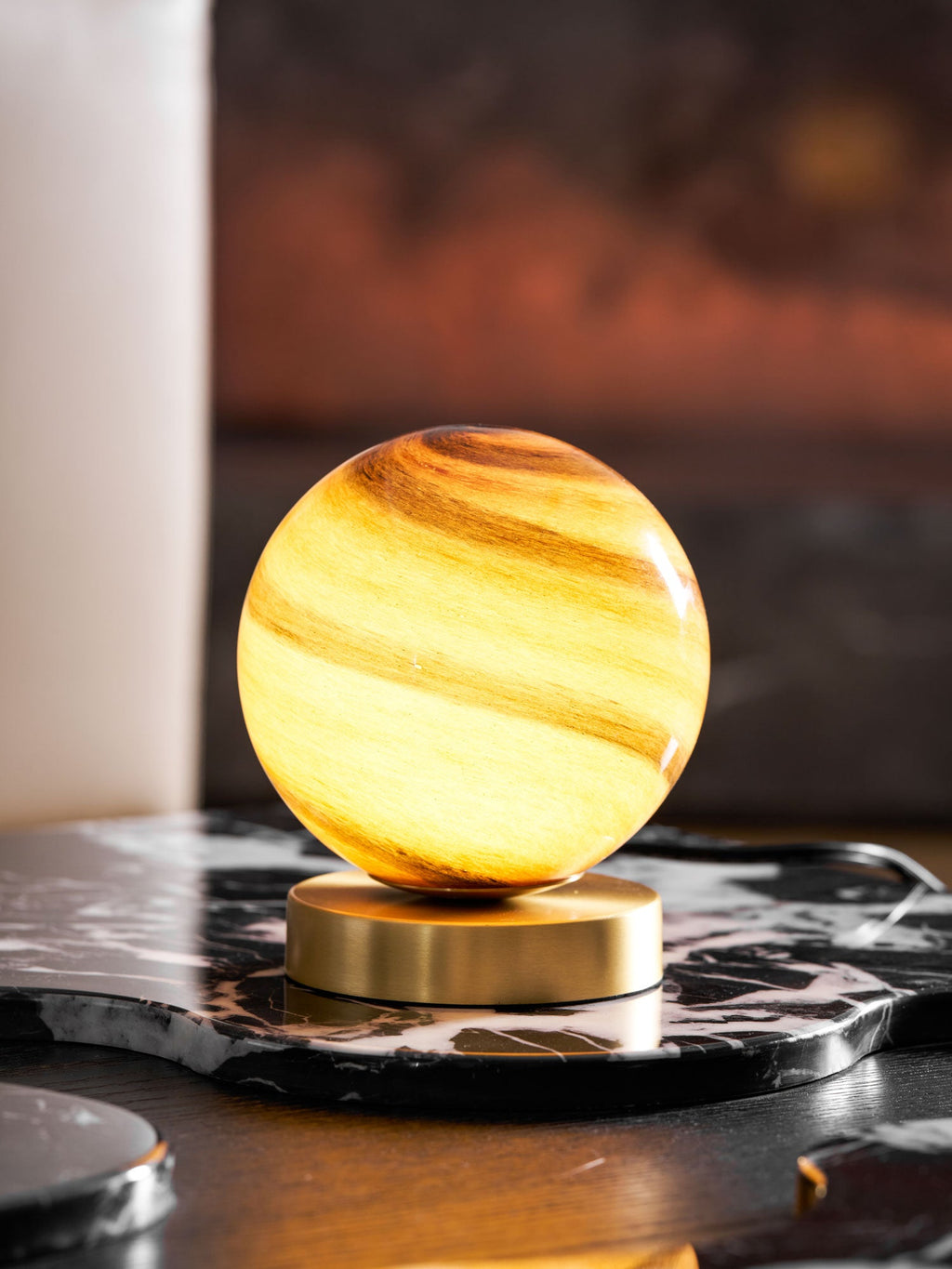 A lit Clérachic globe lamp on black marble, its amber glass glowing in front of a white candle and a glowing red-brown wall.