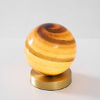 Front view of this Clérachic lamp lit, its banded amber globe resting on a brass disc against a light grey backdrop.