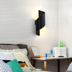 A black Clérachic spiral wall light glows above a bed with a bedside table, a mirror and a small jewellery chest, against white brick.