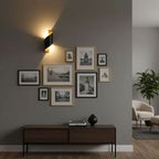 Lit Clérachic wall light on a grey wall above a dark wood sideboard, beside a gallery of black-and-white framed prints.