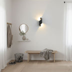 Black Clérachic wall light throwing warm light up and down in a bright hallway with a wooden bench, a round mirror and a shelf.