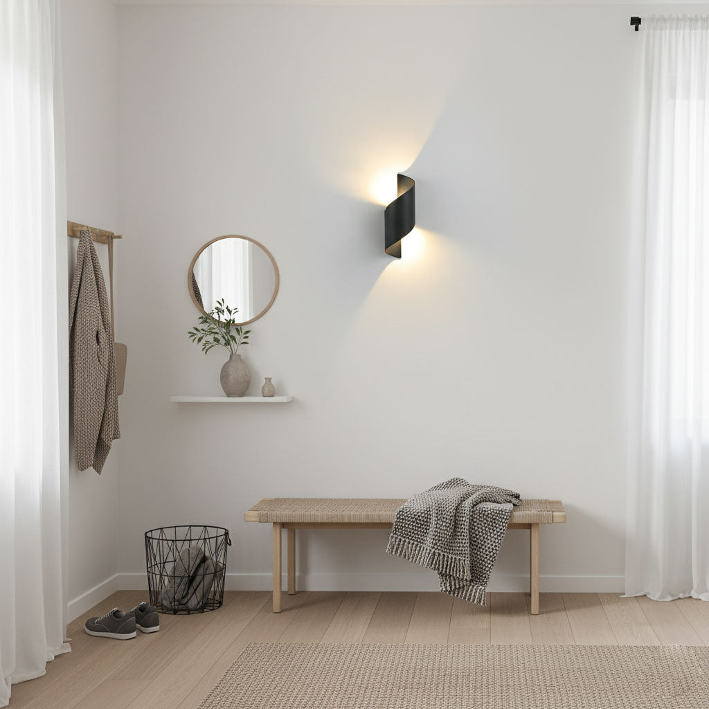 Black Clérachic wall light throwing warm light up and down in a bright hallway with a wooden bench, a round mirror and a shelf.