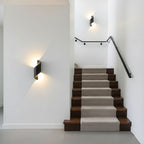 Two black Clérachic wall lights lit along a white stairwell with a dark handrail and carpeted wooden steps.