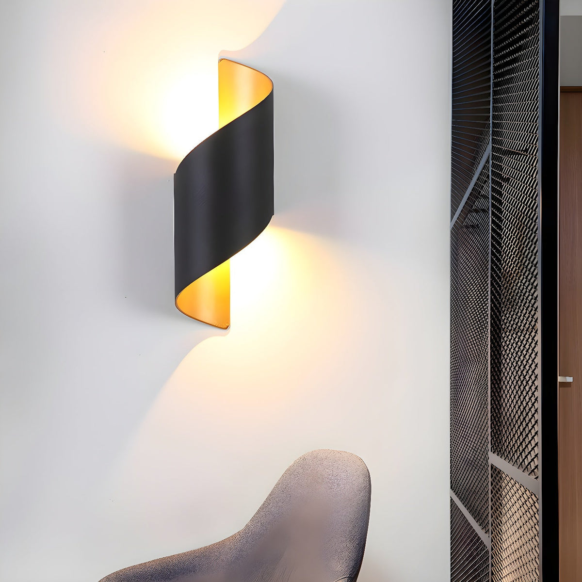 Clérachic spiral wall light in black, lit on a light wall beside a dark mesh screen and the back of a chair.