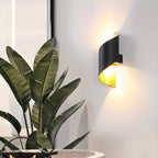 Black Clérachic wall light lit among large green leaves, its gold-toned inside glowing against a pale wall.