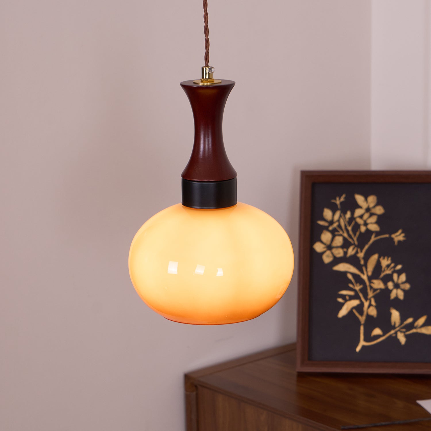 The Clérachic Swirled Amber Glass Pendant Light lit to a warm marigold beside a framed botanical print on a walnut sideboard.