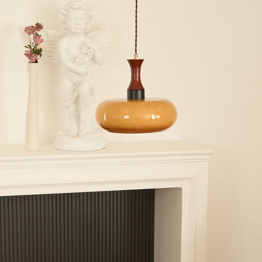A Clérachic Swirled Amber Glass Pendant Light drops over a white fireplace mantel set with a cherub statue and dried roses.