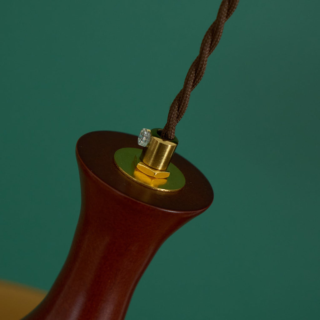 Detail of the brushed brass cord grip and set screw on the walnut neck of a Clérachic amber glass pendant, green backdrop.