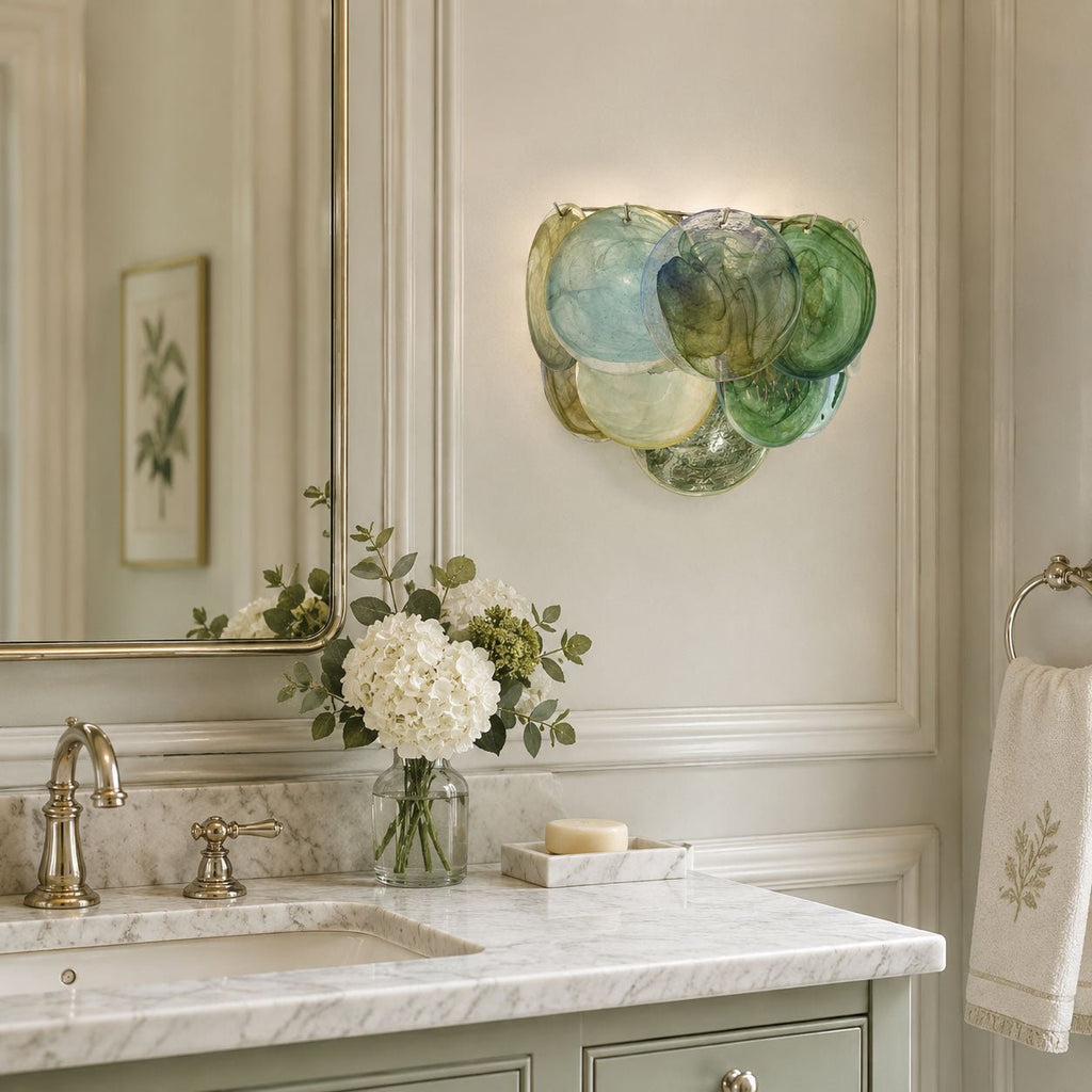 A multicolour disc wall light from Clérachic in blue, green and cream glass, hung beside a bathroom mirror above a marble vanity with white hydrangeas.