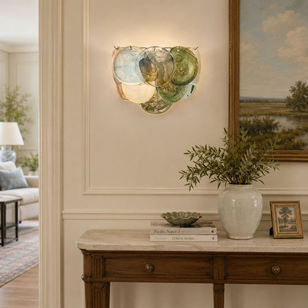 Lit blue and green glass-disc wall sconce from Clérachic above a marble-topped console, with a landscape painting beside it.