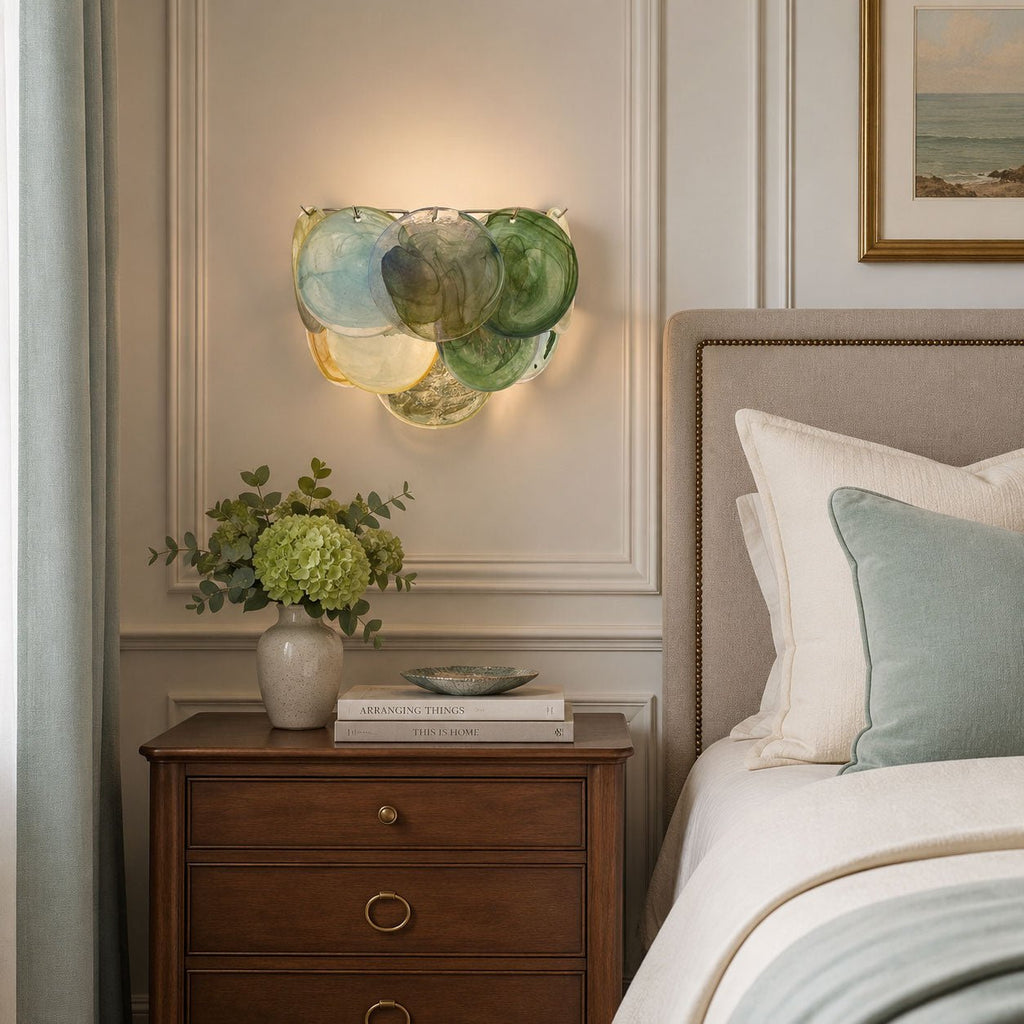 Wall light from Clérachic in blue and green discs glowing above a wooden bedside table, with a green plant and a pale bed.