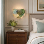 Wall light from Clérachic in blue and green discs glowing above a wooden bedside table, with a green plant and a pale bed.