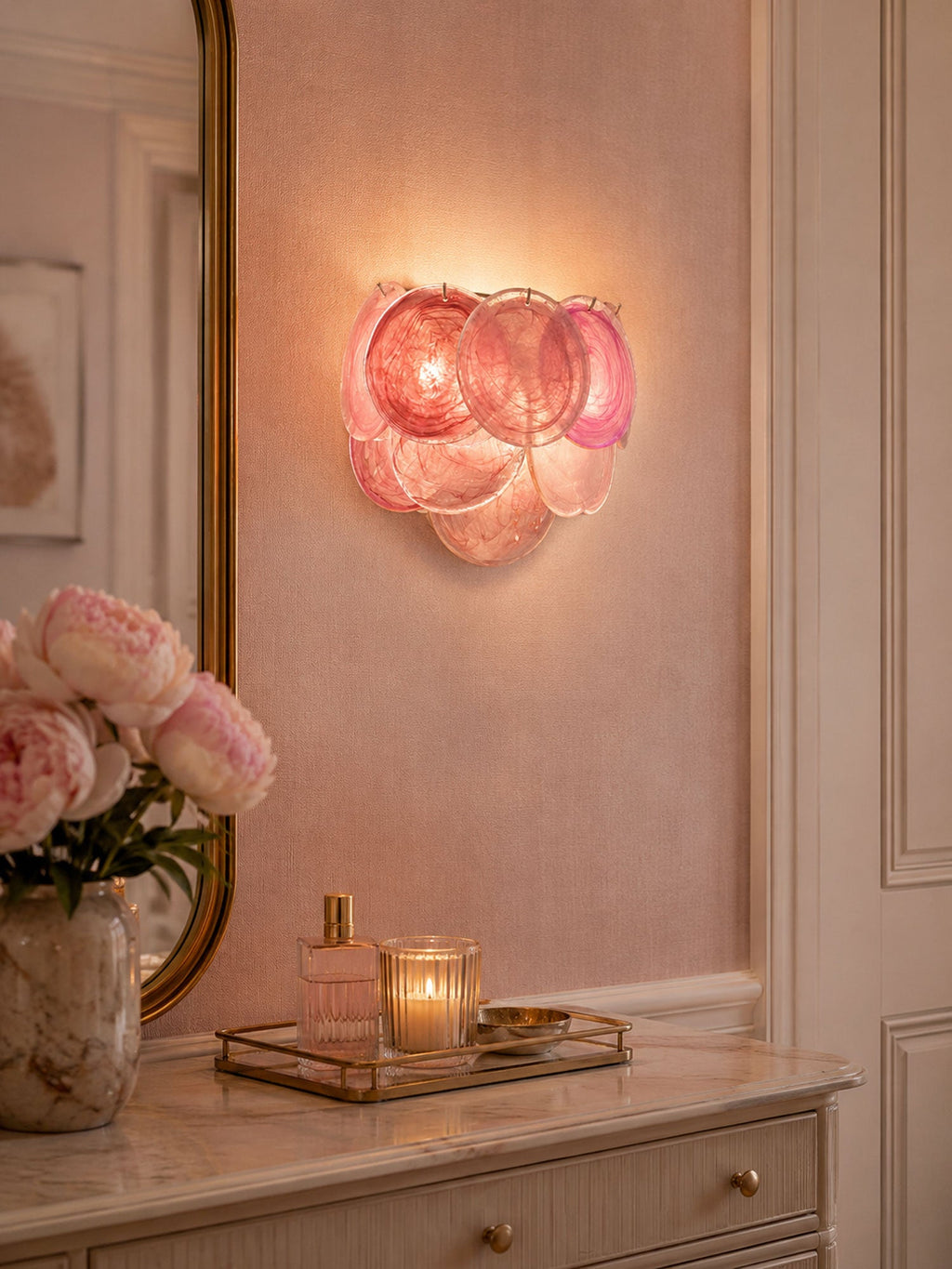 Pink disc wall light from Clérachic lit above a dressing table with a candle, perfume and peonies, against a rose-coloured wall.