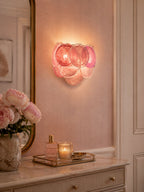 Pink disc wall light from Clérachic lit above a dressing table with a candle, perfume and peonies, against a rose-coloured wall.