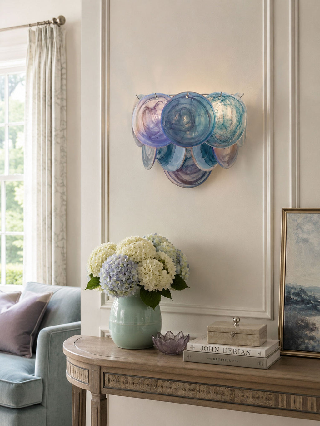 Blue and lilac disc wall light from Clérachic above a carved console table with pale hydrangeas in a green vase.