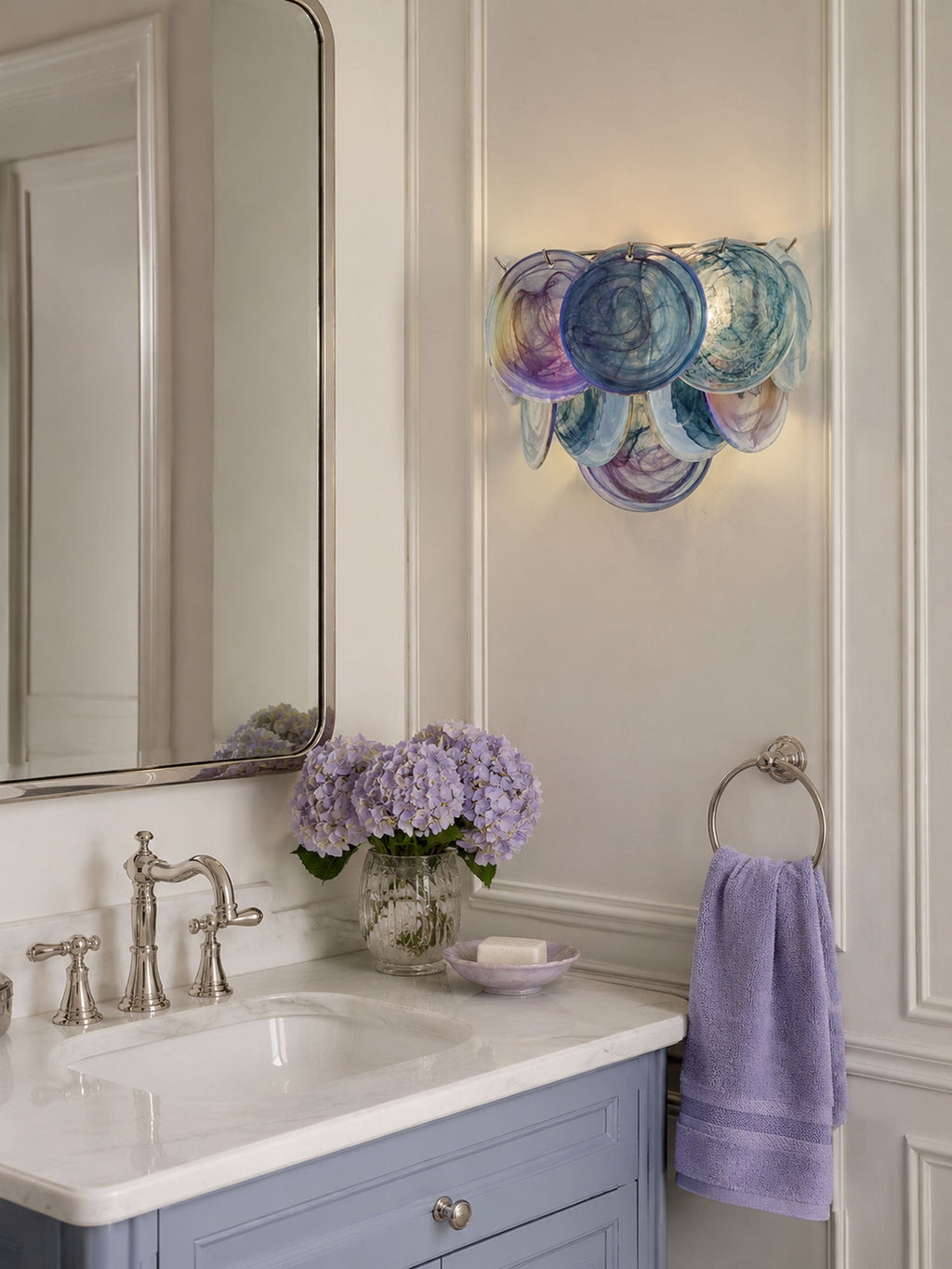 Lit blue-violet disc wall sconce from Clérachic beside a tall mirror above a bathroom basin with lilac flowers and a lavender towel.