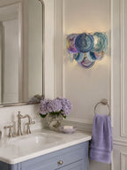 Lit blue-violet disc wall sconce from Clérachic beside a tall mirror above a bathroom basin with lilac flowers and a lavender towel.