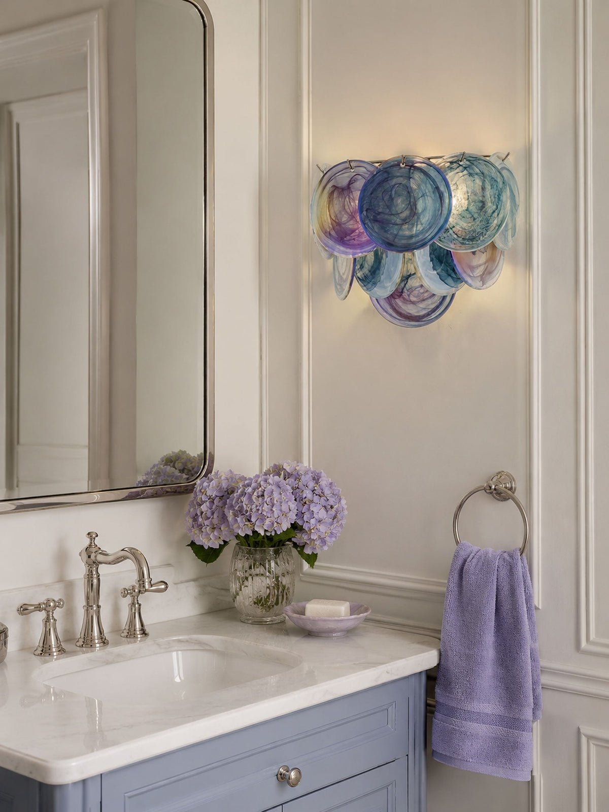 Lit blue-violet disc wall sconce from Clérachic beside a tall mirror above a bathroom basin with lilac flowers and a lavender towel.