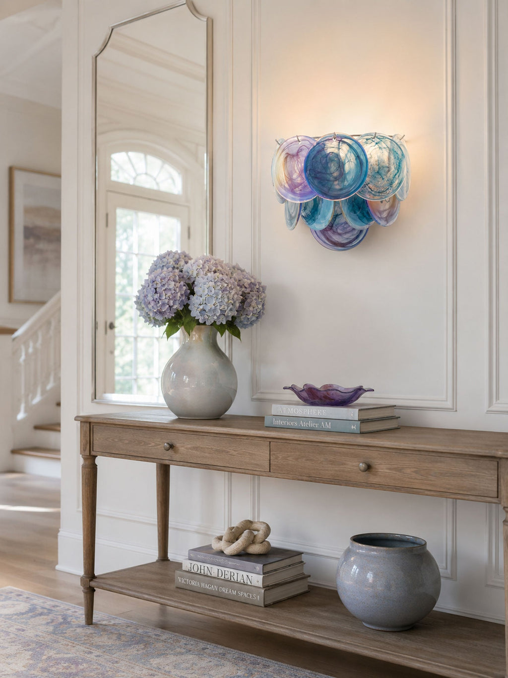 Iridescent blue and violet glass-disc wall light from Clérachic above a wooden console with a hydrangea vase and a grey ceramic pot.