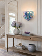 Iridescent blue and violet glass-disc wall light from Clérachic above a wooden console with a hydrangea vase and a grey ceramic pot.